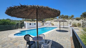 A swimming pool with stone flooring, a small white building, and outdoor seating with a table and chairs.
