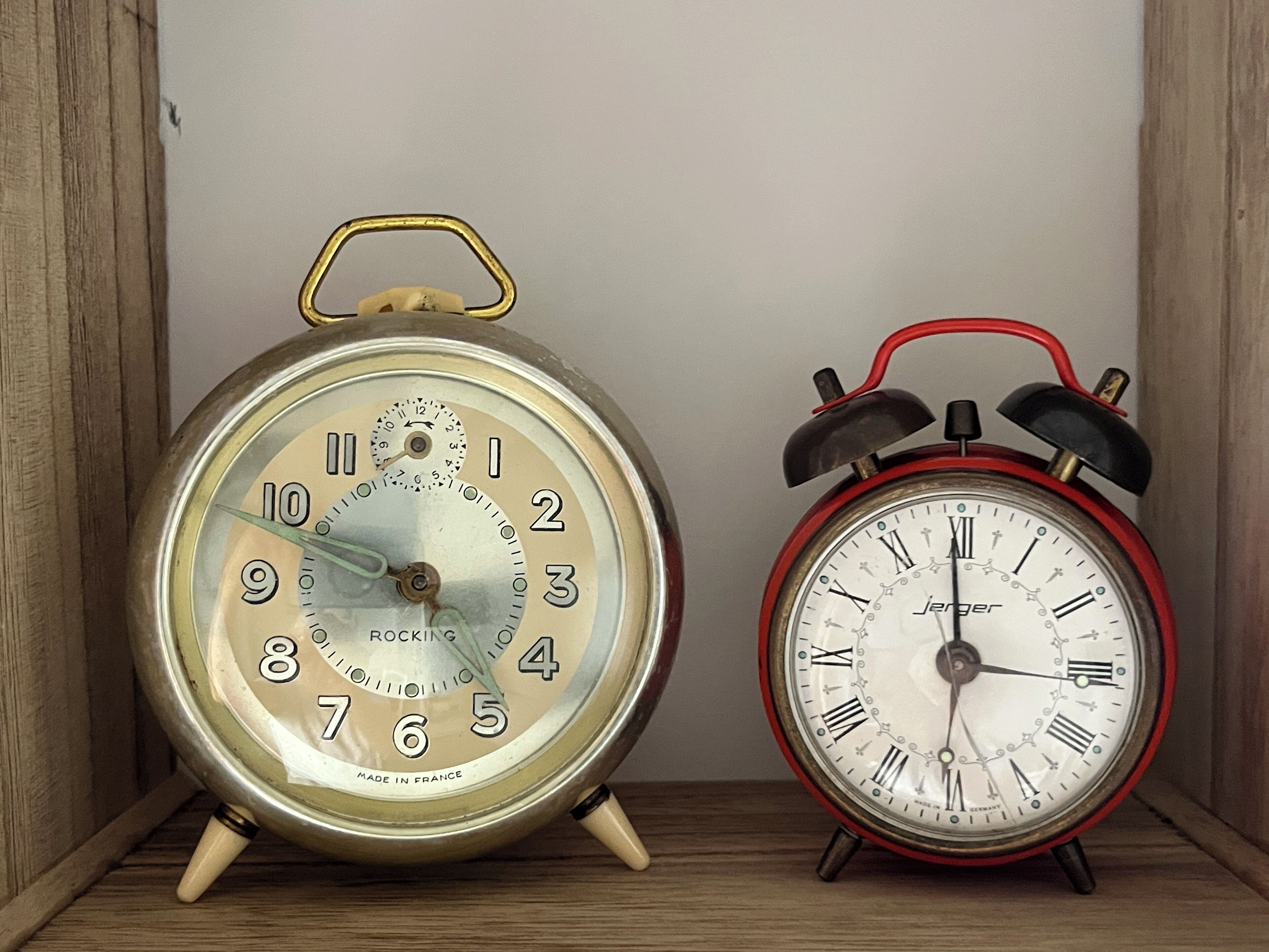 Deux vieux réveils, l’un avec un cadran beige et l’autre rouge, sont posés sur une étagère en bois.