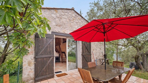 Un patio al aire libre con una mesa y sillas de madera, un paraguas rojo y un edificio de piedra con contraventanas de madera.