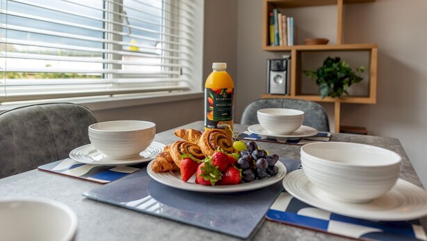 A breakfast setup with croissants, strawberries, grapes, and orange juice on a table with white plates and bowls.