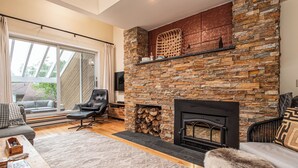 A cozy living room with a stone fireplace, a comfortable sofa, and a large window with curtains. - Mountain condo by Sugarbush w/ fireplace (Warren)