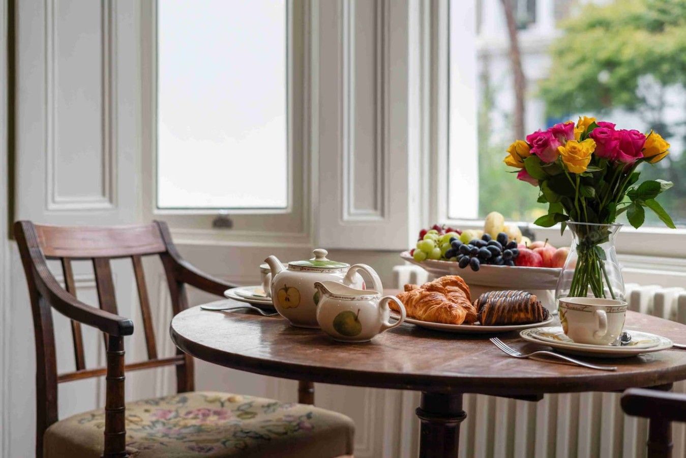 A dining table with a teapot, cups, pastries, and a fruit bowl.