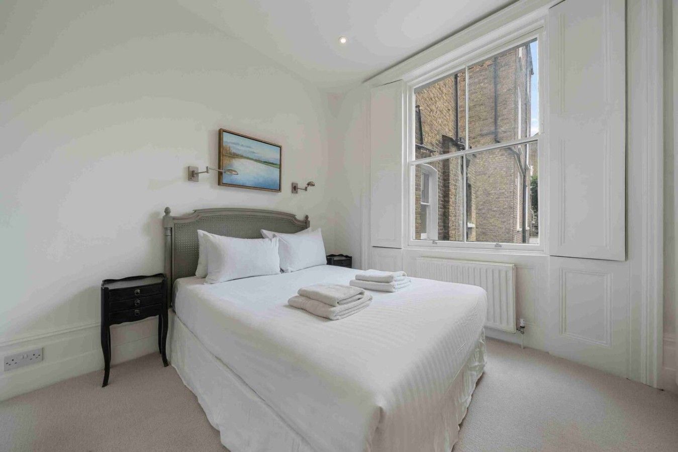 A bedroom with a large window, a bed with white linens, a nightstand, and a painting on the wall.