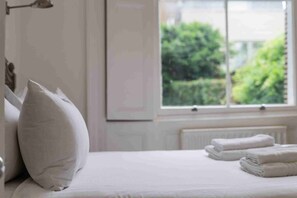 A neatly made bed with pillows and folded towels, a window with a view of greenery, and a radiator under the window.