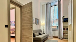 A modern living room with a grey sofa, a floor lamp, a bookshelf, and a large window with curtains. - [Aquarium] Porto Antico Vanilla Apartment (Genova)