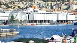 A waterfront marina with boats docked, a large glass greenhouse structure, and a cityscape in the background. - [Aquarium] Porto Antico Vanilla Apartment (Genova)