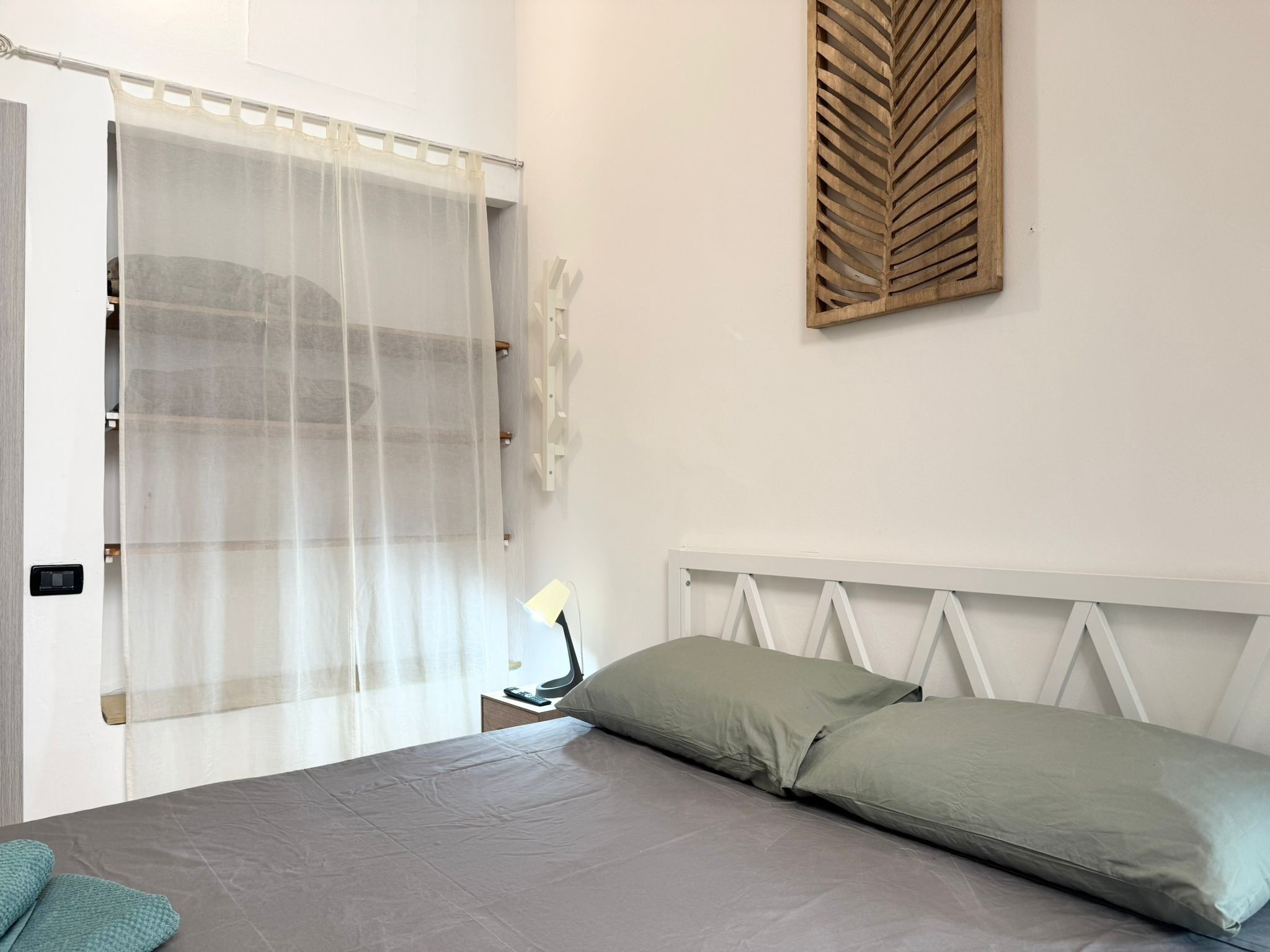 Ein Schlafzimmer mit einem Bett, einem Nachttisch mit Lampe, einem transparenten Vorhang und einer Holzdeko an der Wand.