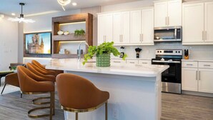 A modern kitchen with white cabinets, a central island with bar stools, a microwave, an oven, and a decorative plant. - Spacious 6-bedroom house in Reunion - Fully themed game room - xbox and more (Kissimmee)