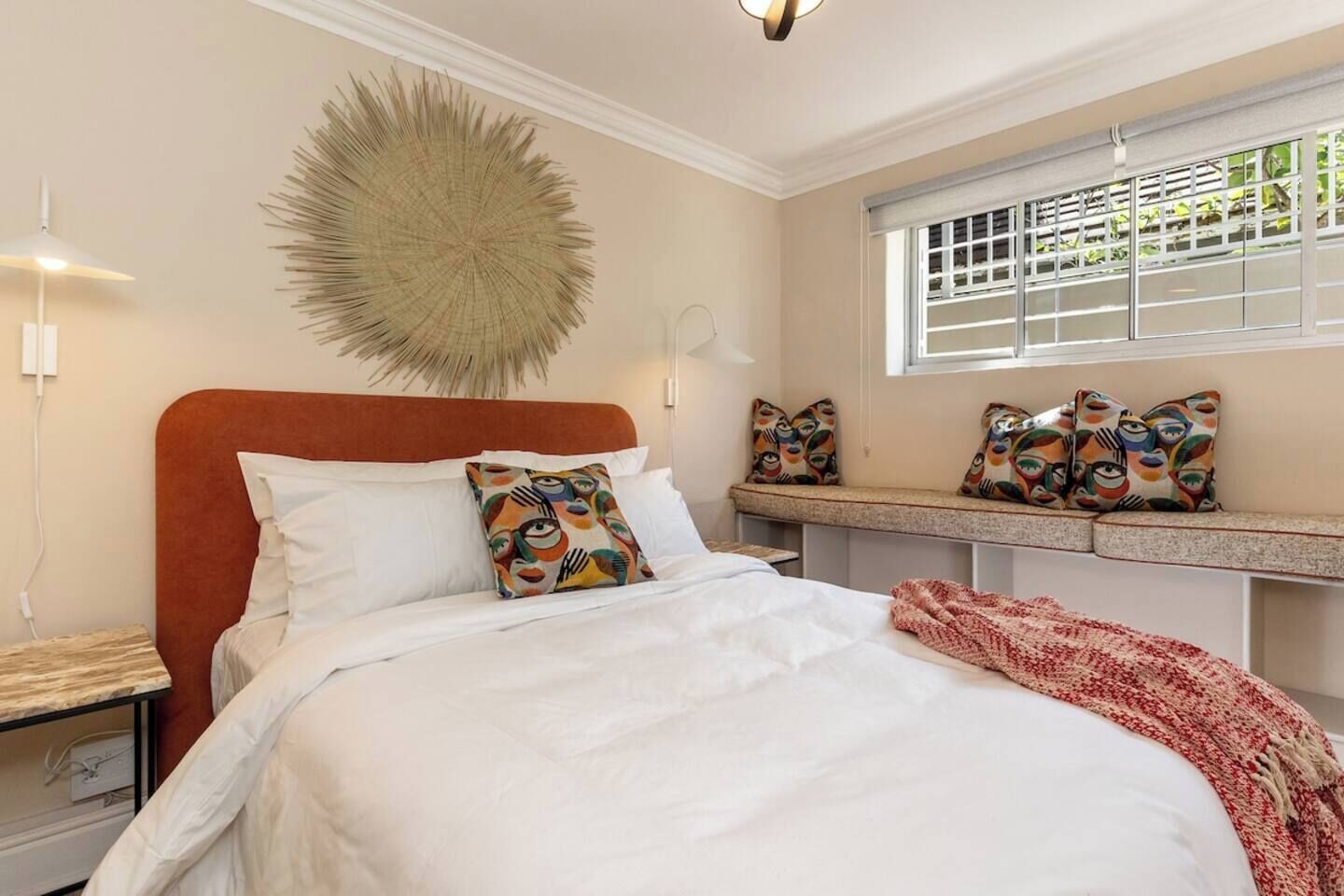 A bedroom with a bed, a headboard, a window with blinds, a bedside table, and a wall-mounted lamp.