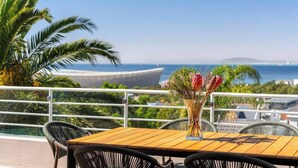 A terrace with a wooden table and chairs, a vase with flowers, and a view of the sea and distant land. - Your CT experience starts here (Cape Town)