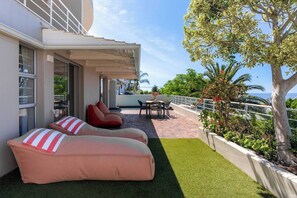 A balcony with a sofa, chairs, and a table, overlooking a lush green area and a tree.
