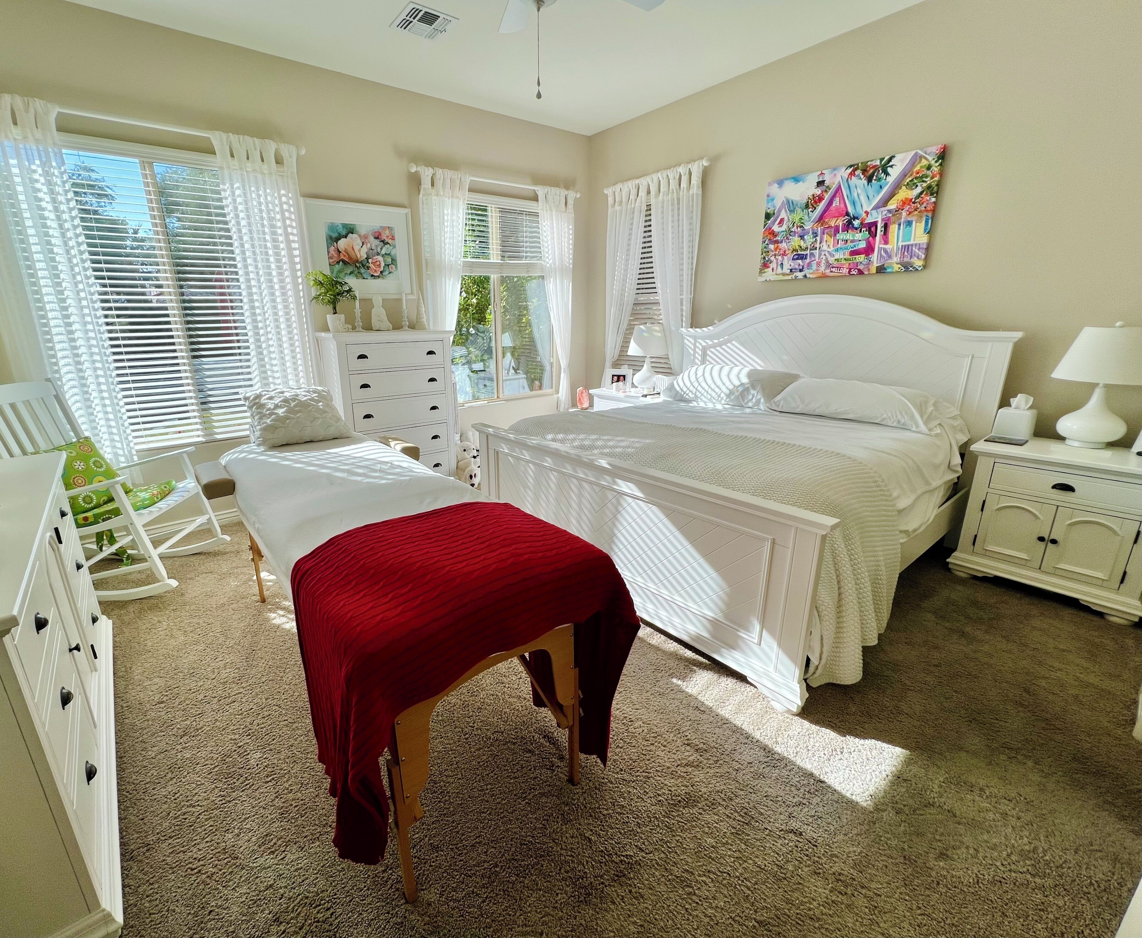 A bedroom with a large bed, a bedside table, a rocking chair, a dresser, and a window with blinds.