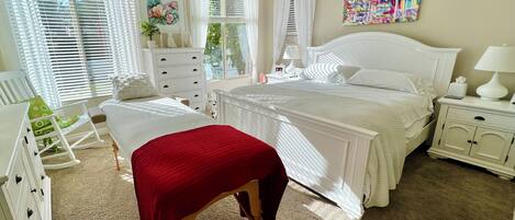 A bedroom with a large bed, a bedside table, a rocking chair, a dresser, and a window with blinds.