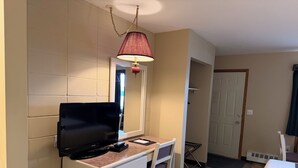 Ein Hotelzimmer mit Flachbildfernseher, Schreibtisch mit Stuhl, Pendelleuchte, Tür und Bett.