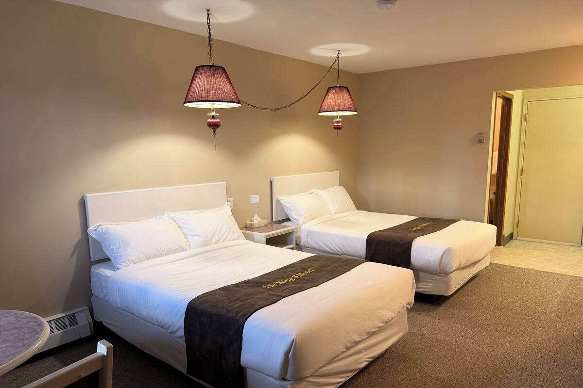 Quarto de hotel com duas camas, uma mesa de cabeceira, uma luminária e uma porta.
