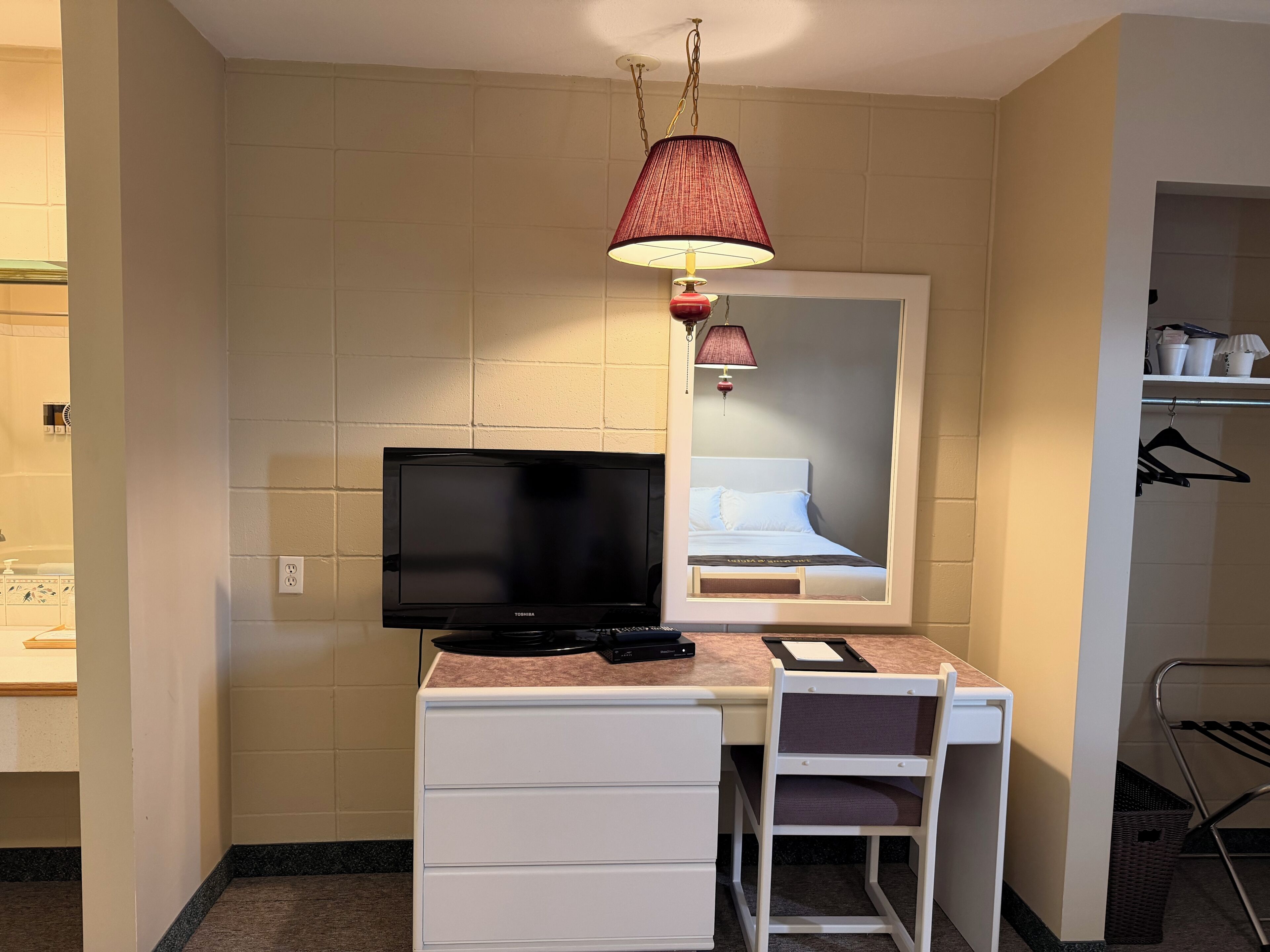 Camera d'albergo con televisione a schermo piatto, una scrivania con cassetti, una sedia, una lampada a sospensione e uno specchio sopra il letto.