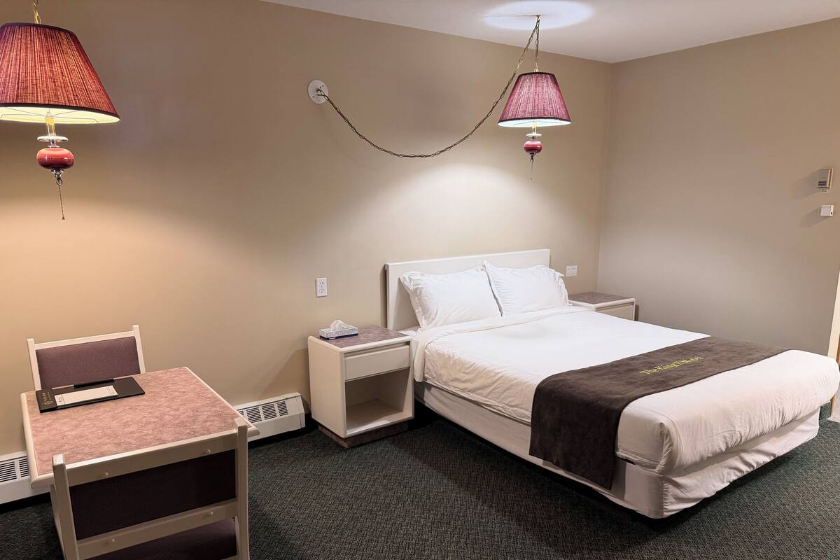 Quarto de hotel com uma cama, uma escrivaninha, dois criado-mudos, uma cadeira e duas luminárias fixadas na parede.