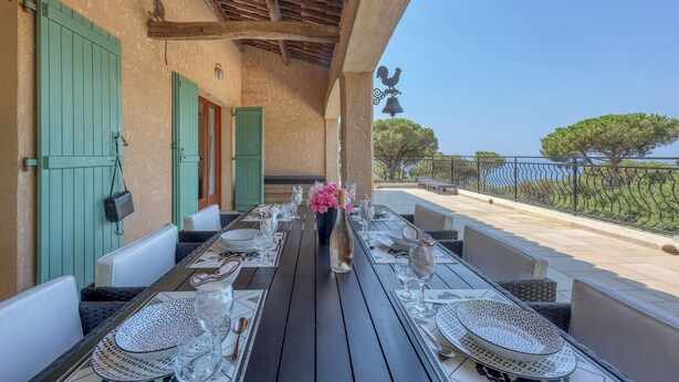 An outdoor dining area with a long table set for two, surrounded by chairs, a vase with flowers, and a view of the sea.