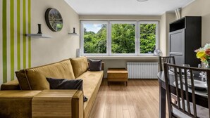 A modern living room with a sofa, a dining table with chairs, a TV, and a large window with a view of greenery.
