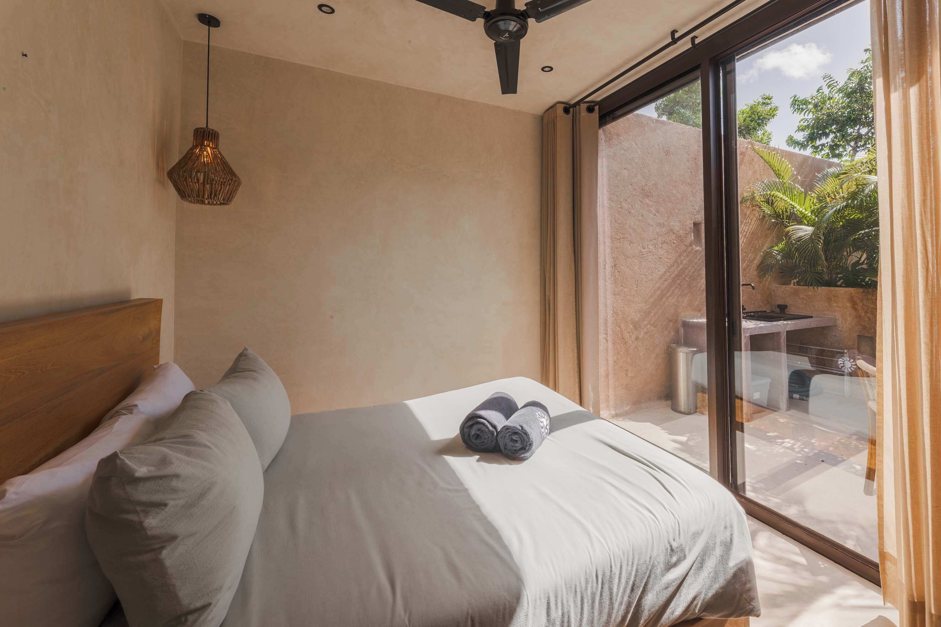 A bedroom with a bed, pillows, a ceiling fan, a hanging light fixture, and a view through a glass door.
