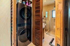 A laundry area with two front-loading washing machines, a wooden slatted door, and a glass shower enclosure. - Serene 2BR Apartment with Private Pool, Kokoon Pueblo II (Tulum)