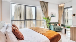 Een moderne hotelkamer met een groot bed, een groene bank, een kleine tafel en uitzicht op hoge gebouwen door grote ramen.