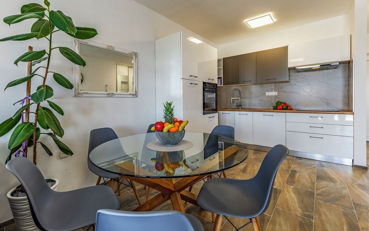 Une cuisine moderne avec des meubles blancs, un pare-feu en marbre et un coin repas comprenant une table en verre et des chaises bleues.
