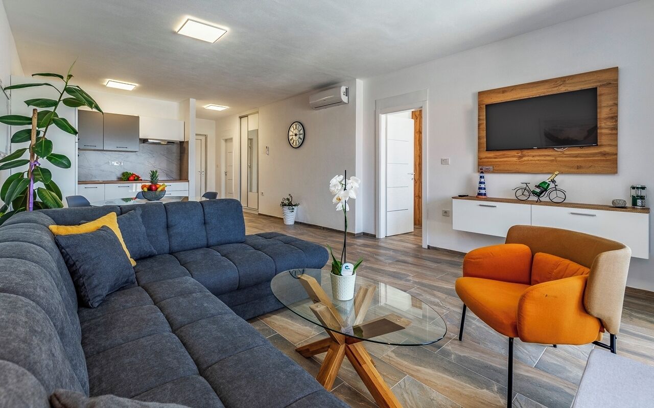 A modern living room with a sectional sofa, a glass coffee table, and an orange armchair. A TV is mounted on the wall, and there is a kitchen in the background.