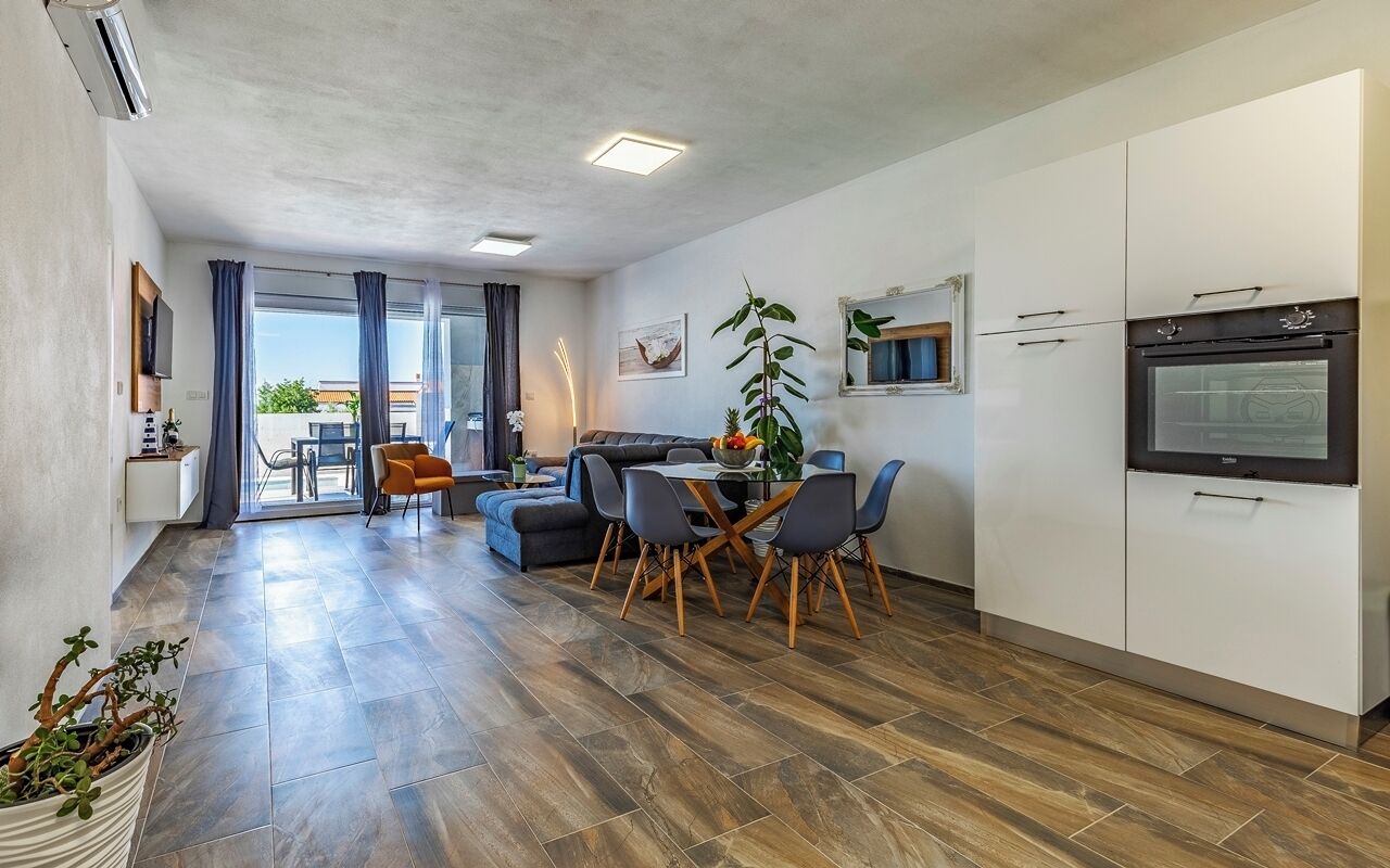 Una sala de estar moderna con un sofá, una mesa de comedor y sillas. Hay una zona de cocina con armarios y un horno. Una planta está sobre la mesa de comedor.