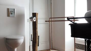 A bathroom with a white toilet, a wooden stool, a metal rack, and a window with natural light.