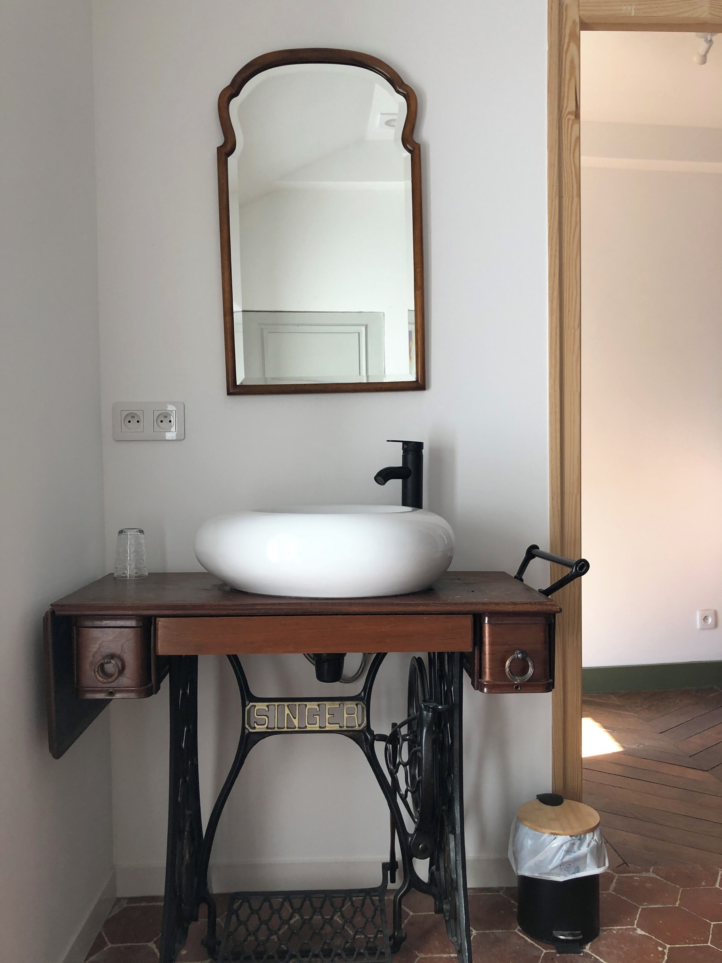 Un baño con un lavamanos sobre la base antigua de una Singer, un lavabo blanco y un espejo con marco de madera.