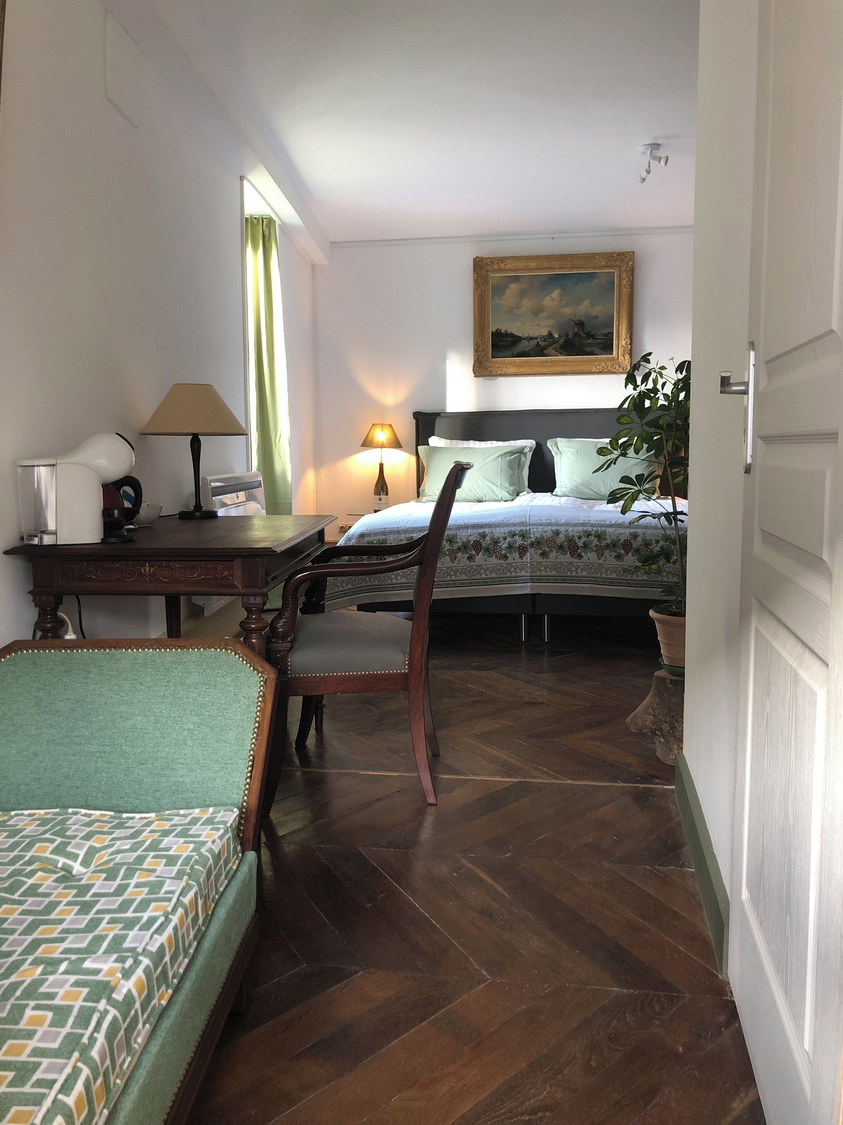 A hotel room with a bed, a desk with a lamp, a chair, and a potted plant.
