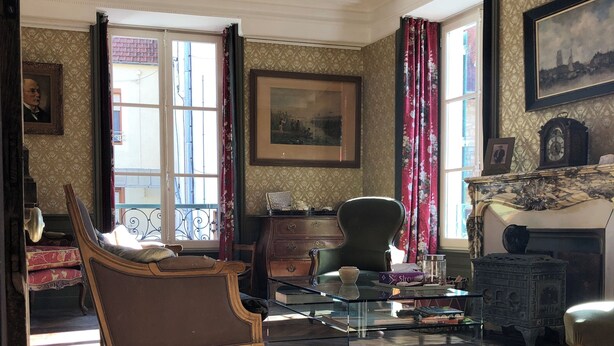 Una habitación con suelo de madera, un ventanal con cortinas, un cuadro en la pared y una chimenea con repisa.