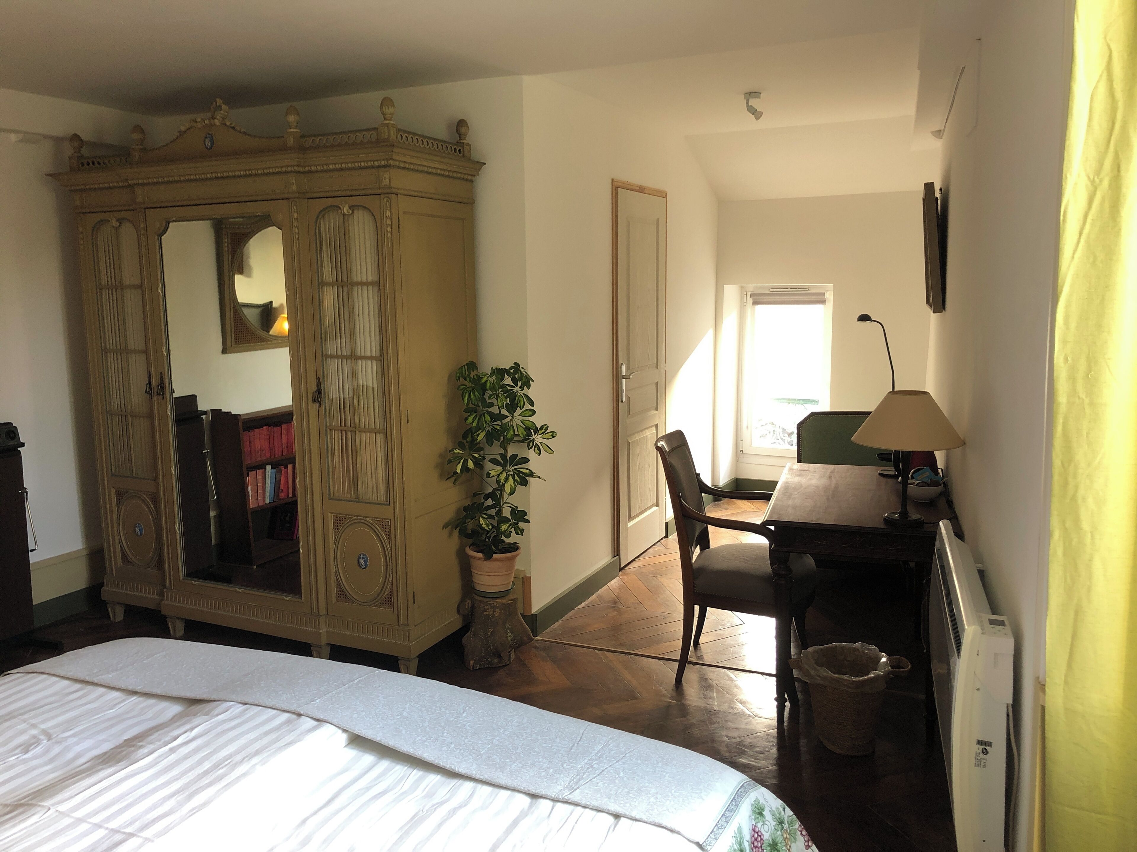 Une chambre avec un grand lit, une armoire, un bureau avec une lampe, une plante en pot et une fenêtre laissant entrer la lumière naturelle.