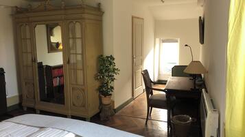 Une chambre avec un grand lit, une armoire, un bureau avec une lampe, une plante en pot et une fenêtre laissant entrer la lumière naturelle.
