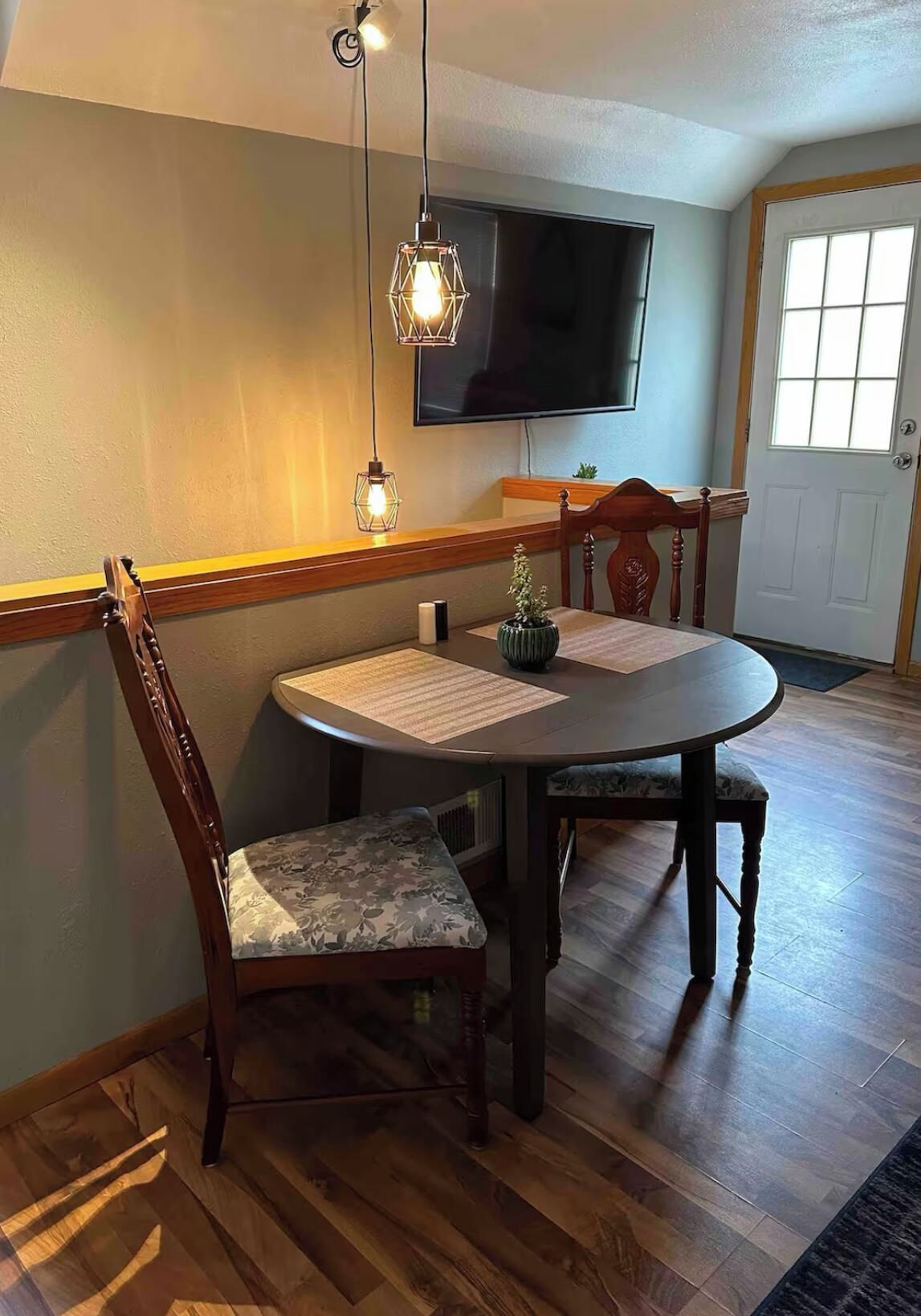 Un comedor con una mesa redonda, dos sillas, un televisor montado en la pared y una puerta con ventana.