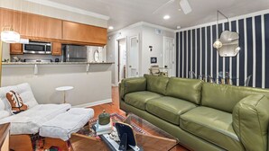 A living room with a green leather sofa, a white couch, and a coffee table with books and decorative items. There is a kitchen area with wooden cabinets and a microwave. - 
Travis Heights Modern Getaway - Corporate Housing Rental (Austin)