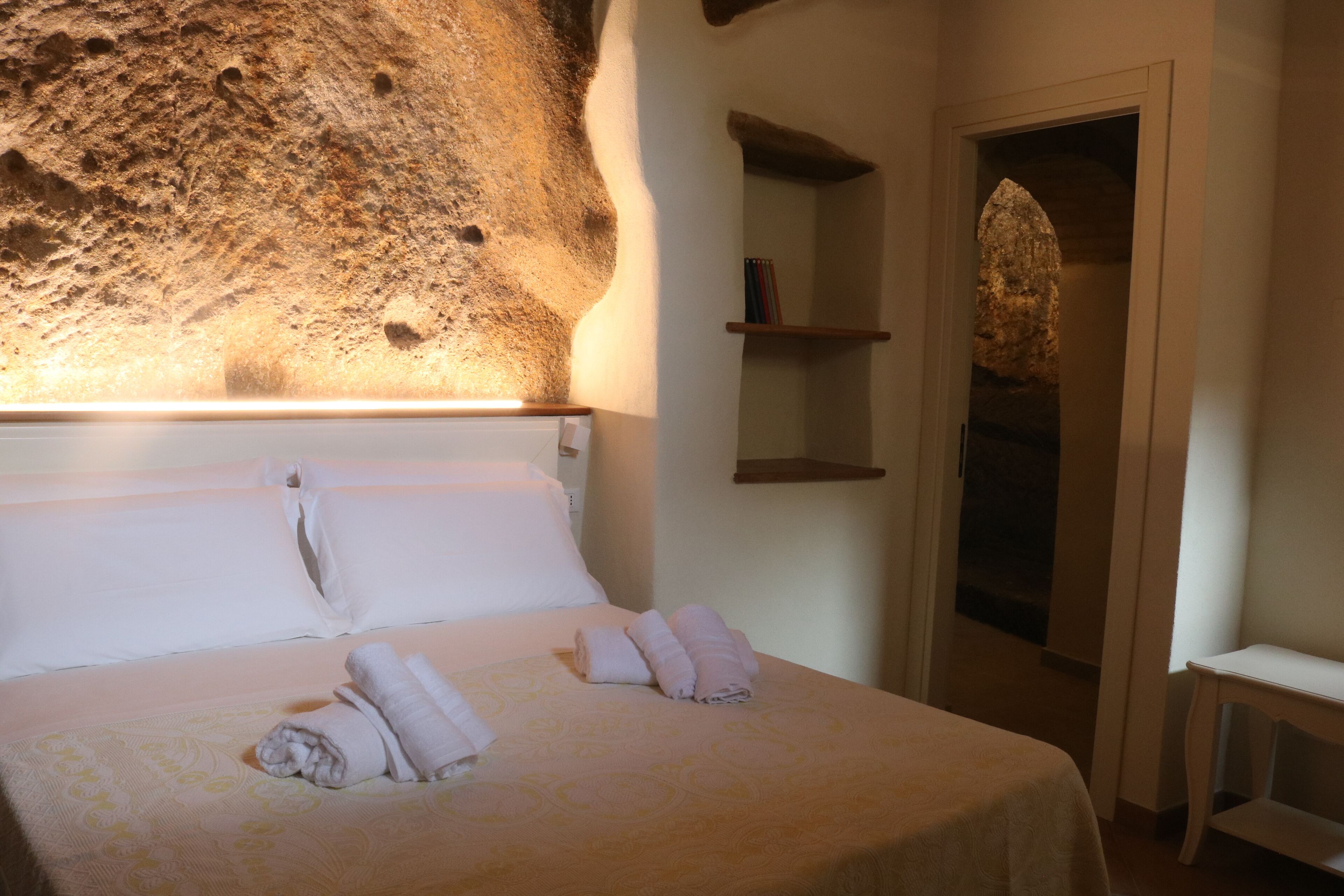 Ein Hotelzimmer mit einem großen Bett, ordentlich gefalteten Handtüchern, einer Steinwand und einem kleinen Regal mit Büchern.