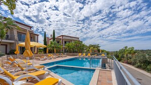 A pool area with sun loungers and umbrellas, a clear sky, and a building with balconies.