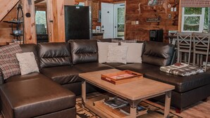 A cozy living room with a brown leather sectional sofa, a wooden coffee table, and a kitchen area visible through an open door.