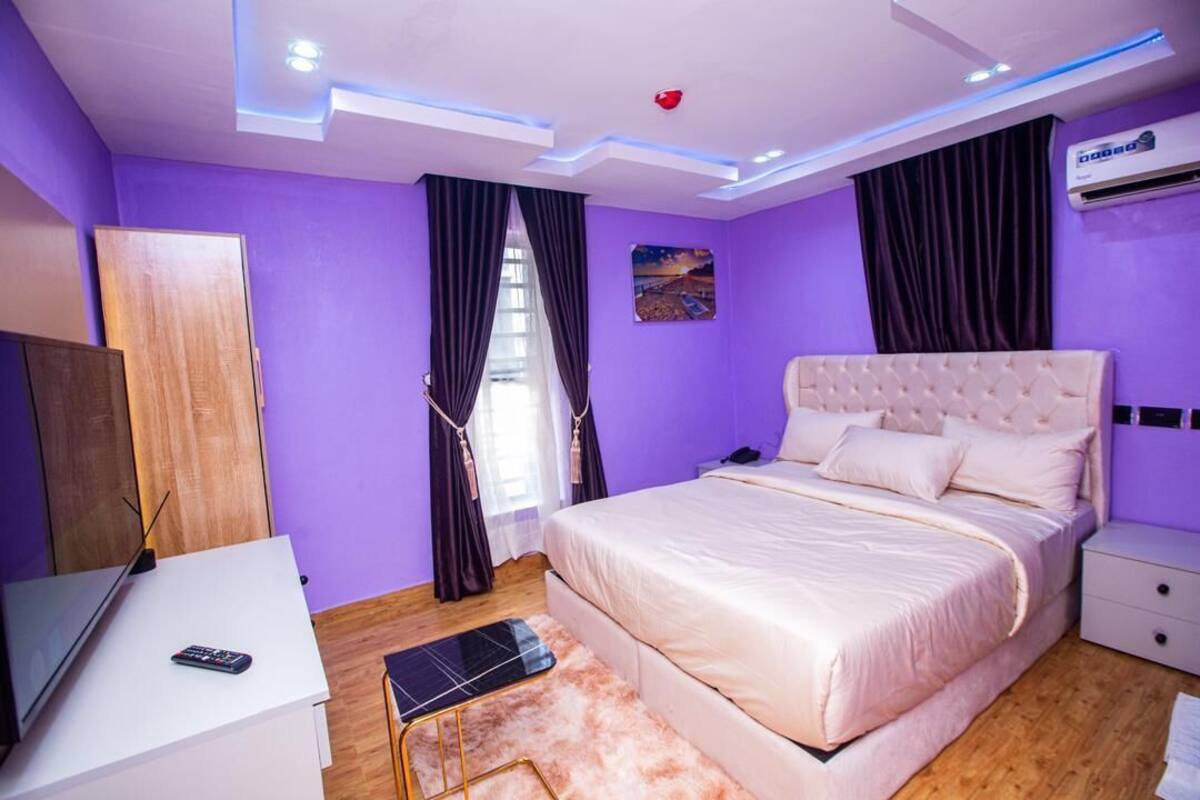 Quarto moderno com uma cama grande, uma mesinha de cabeceira, uma televisão e uma janela com cortinas.