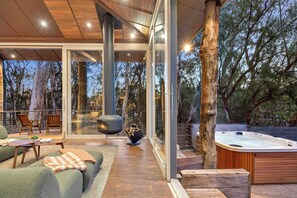 A modern outdoor living area with a hot tub, seating, and glass walls offering views of the surrounding forest.
