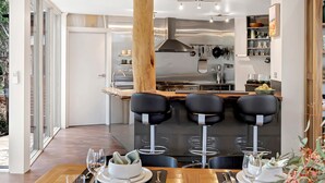 A modern kitchen with a central island, bar stools, and a dining area with a table set for two. - The Treehouse (Moama)