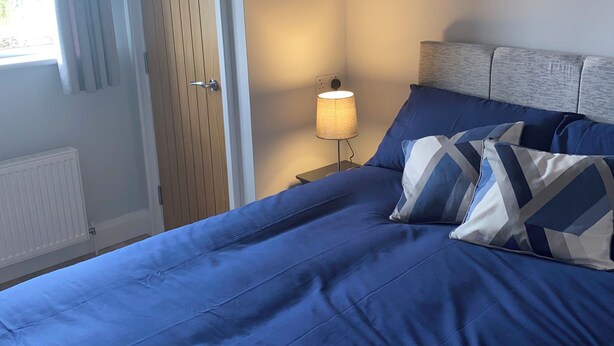 A bedroom with a bed, a nightstand, a lamp, a window with curtains, and a door.