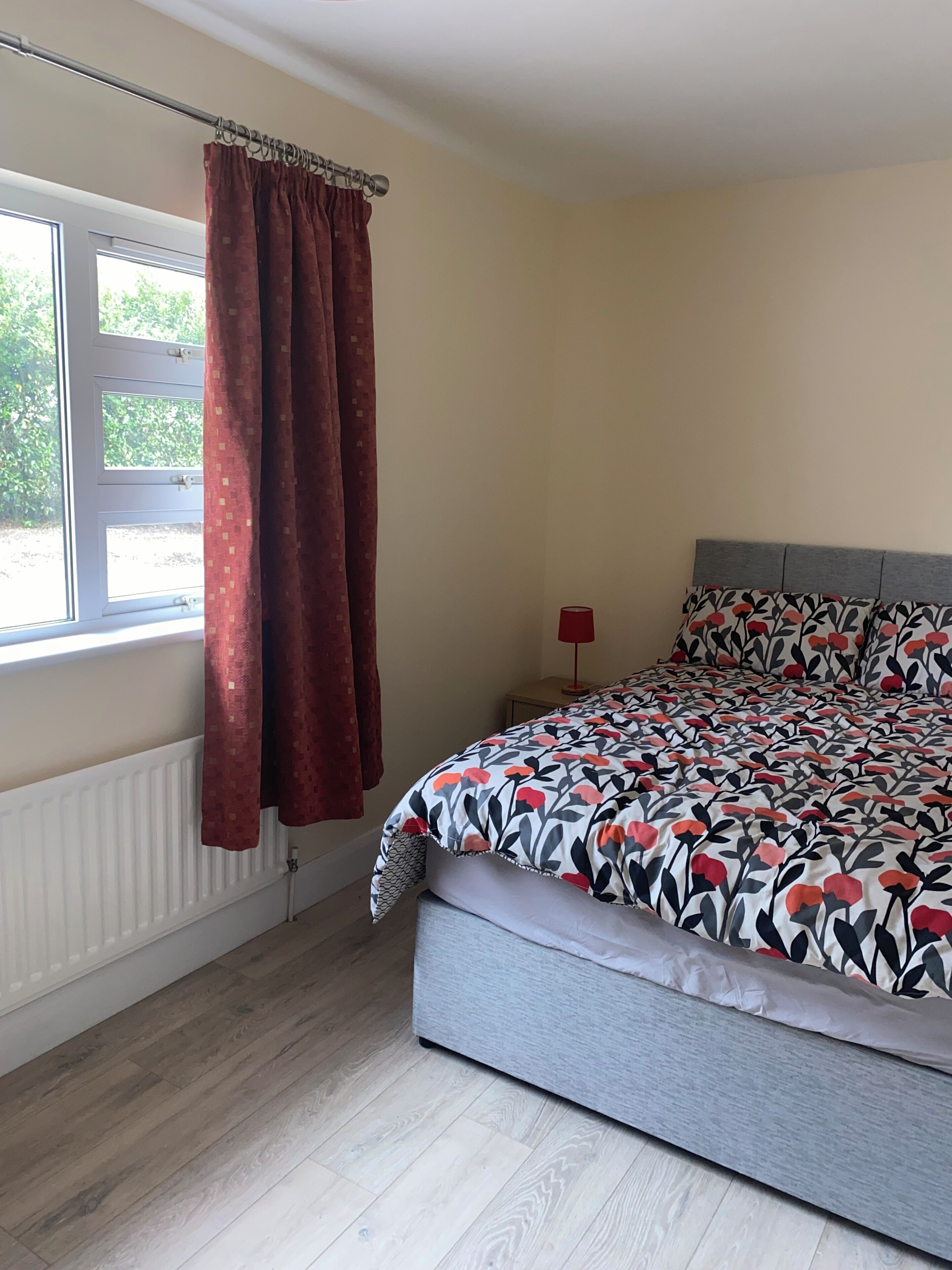 A bedroom with a bed, a window with curtains, a radiator, and a bedside table with a lamp.