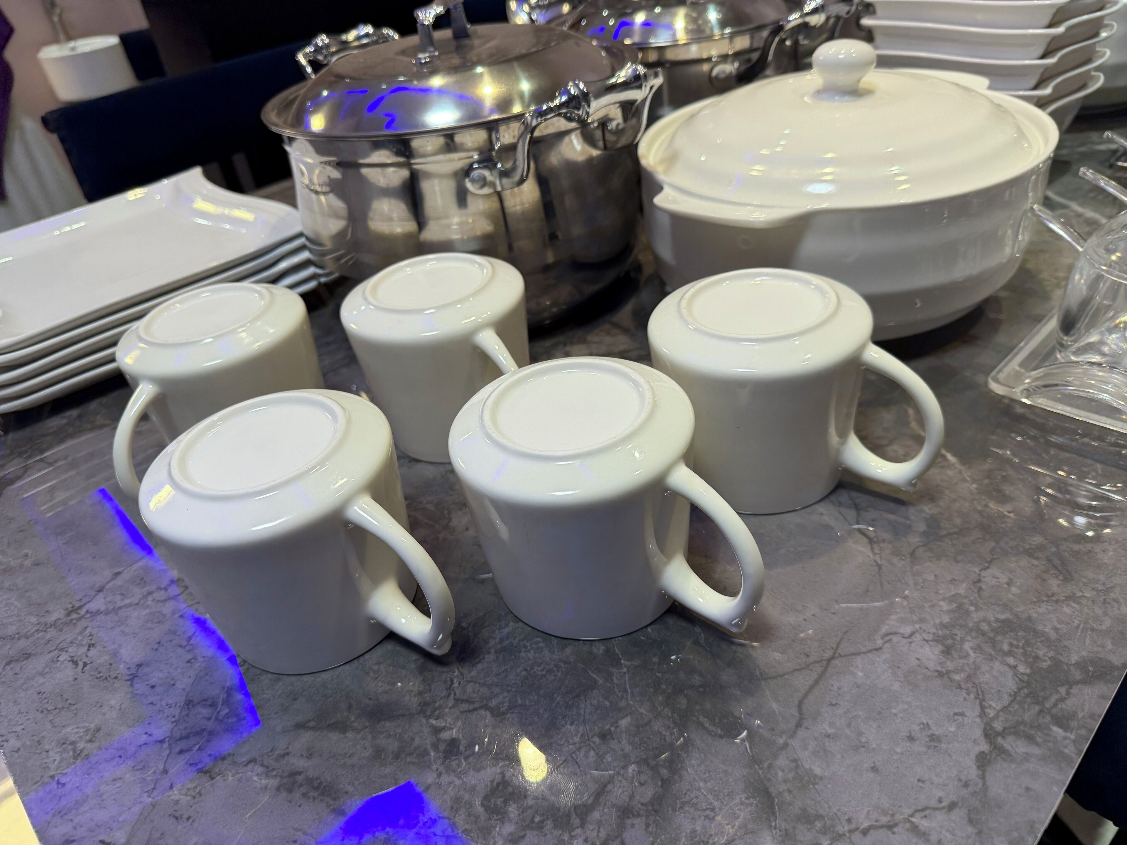 Une table dressée avec des théières, des tasses et des assiettes en céramique blanche, une casserole en acier inoxydable et un grand bol blanc en arrière-plan.