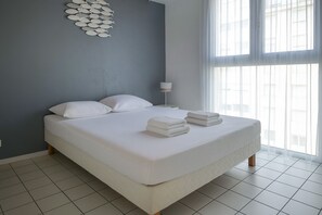 Ein ordentlich bezogenes Bett mit weißen Leinen und Kissen, eine Wandleuchte und ein großes Fenster mit transparenten Vorhängen.