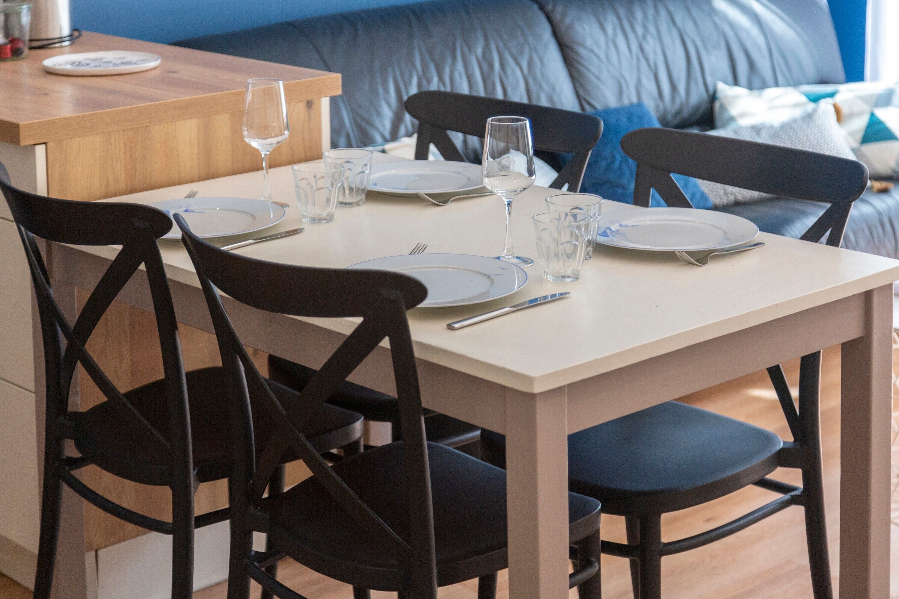 Une table à manger est dressée avec des assiettes, des couverts et des verres, entourée de chaises noires dans une pièce où l’on aperçoit un canapé bleu à l’arrière-plan.
