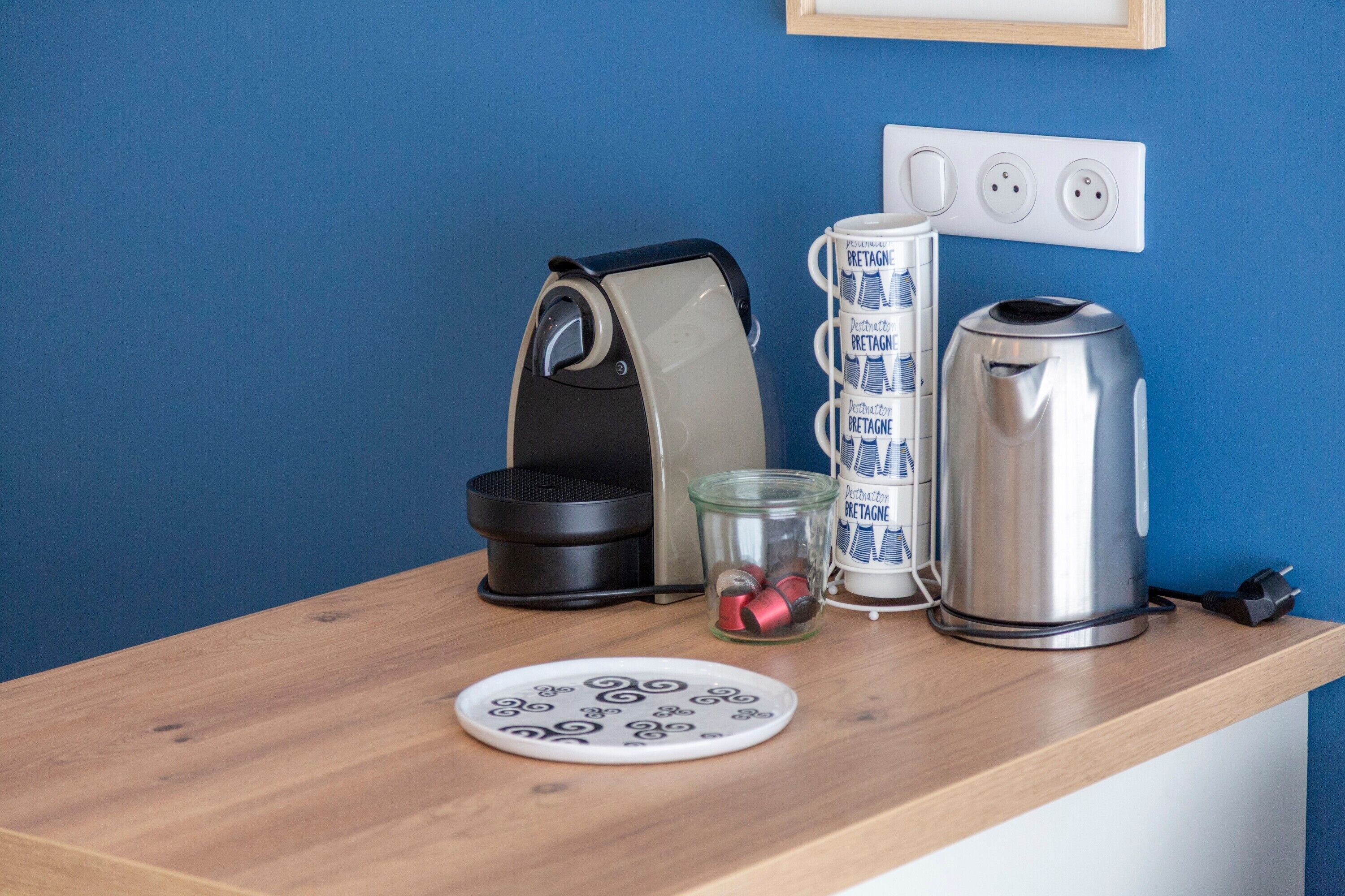 Un plan de travail de cuisine avec une machine à café, une bouilloire, un bocal en verre contenant des sachets de thé et une assiette décorée.