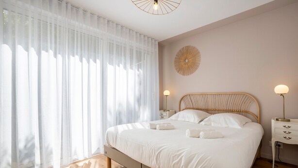 A bedroom with a bed, bedside tables, a wall clock, and sheer curtains.